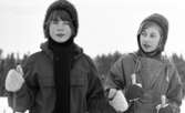 Hästnäs, 20 januari 1966

På bilden i förgrunden syns det två flickor som är klädda i vinterkläder. Flickorna har stickade mössor på sina huvuden. Flickan åt vänster har stickade vantar på sina händer och en lång stickad skarf runt sin hals som hänger ner. Båda flickor håller skidstavar i sina händer.