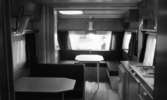 Husvagnar, 26 januari 1966

I bildens förgrund syns en husvagnsinteriör, med diskmaskin, spis, skåp, lådor och köksbänk. Runt matsalsbordet vid fönstret rakt fram i bildens bakgrund finns en soffa. Till vänster finns ytterligare ett bord.