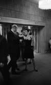 Premiär Julie, 28 februari 1966

En kvinna i klänning samt broderad kofta samtalar med kostymklädd herre inne på teatern under pausen. Andra personer skymtar i bakgrunden.