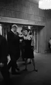 Premiär Julie, 28 februari 1966

En kvinna i klänning samt broderad kofta samtalar med kostymklädd herre inne på teatern under pausen. Andra personer skymtar i bakgrunden.