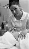 Skönhetsvård, 3 mars 1966

Inne i en skönhetssalong ägnar sig en yrkeskvinna eller kosmetolog åt skönhetsvård av en kunds ansikte. Hon är i färd att måla kundens läppar. Kosmetologen är klädd i vit arbetsklänning med en brosch på, har uppsatt hår i knut, bär örhängen och har målade ögonbryn. På sin vänstra handled har hon ett armbandsur. Kunden ligger på en brits och har håret invirat i en handduk. I bakgrunden skymtar ett bord fyllt med flaskor för skönhetsvård.