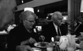 Pensionärslunchen, 3 mars 1966

På bilden centralt syns det en äldre dam och en äldre herre vid ett matbord på en restaurang. Damen har glasögon på sig och hon är klädd i klänning med en brosch i form av en blomma på. Herren bredvid henne är klädd i en mörk kostym, en ljus skjorta med slips. Han har en borstliknande mustasch. Ytterligare en herre sitter till höger i bild. I bakgrunden skymtar serveringsdisken med glas, koppar och fat i montrar.
På bordet ligger en rutig duk. Där finns också tillbringare med mjölk, glas med mjölk i samt tallrikar fyllda med mat, knivar och gafflar. Pensionärerna är i full färd med att äta.