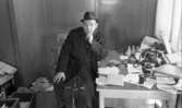 Skofabriksnedläggelse 18 februari 1966
På bilden syns det en man som sitter på en kontorsstol och stödjer sin vänstra armbåge mot bordet bredvid. På bordet står en askkopp, en ask med tändstickor, flera kuvert, en telefonapparat, en kartong med skor, en pärm samt papper. Mannen är klädd i en rock, under rocken har mannen en mörk kostym, en ljus skjorta och en mörk slips. Han har också en hatt på sitt huvud och skor på sina fötter. På golvet vid väggen skymtar ytterligare det pärmar, papper etc.