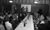 ABF - Norrbysektion, Klänningssömmerskor 21 april 1966

I förgrunden syns ett långbord med vit duk. Ovanpå duken finns kaffekoppar med assietter och skedar till. Vaser med blommor i står även på bordet. Vid bordet sitter damer och herrar. Runtomkring finns andra dukade bord med folk som sitter invid dem. I bakgrunden syns en scen med två sångerskor samt två musiker på framför två mikrofoner. De två sångerskorna är klädda i knäkorta klänningar och de två musicerande herrarna har kavajer, byxor och slipsar på. En av herrarna spelar dragspel och den andre spelar gitarr. Bakom artisterna finns en vit filmduk.