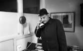 Alltjänst, 7 april 1966

En man i svart ytterrock och hatt talar i en vit kobratelefon inne på ett kontor. Till vänster om mannen hänger en klocka på en vägg. Bakom honom hänger en tavla på en annan vägg.