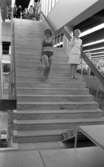 Baddräkter 12 maj 1966

En fotomodell i mönstrad bikini samt med hatt på huvudet går nerför en trappa. I närheten av henne står en kvinnlig konferencier iklädd vit jacka, vit kjol samt vita skor och talar i en mikrofon.
















































































































or. Han går nedför en kort trappa utomhus.