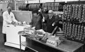 Orubricerat 18 februari 1966

I en charkuterifabrik står två män och en kvinna. Den äldre herrn till höger på bilden håller en bukett blommor i handen. Frysboxen flankeras av en man och en kvinna. Mannen håller i två stycken paket  med påskriften 