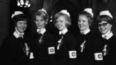 Nya sköterskor, 21 februari 1966

Fem sköterskor klädda i sina yrkesuniformer. På sina vänsterarmar har de armbindlar med kors på. Broscher med kors är fästade i mitten av kragarna framtill och på uniformsklänningarna på vänstersidan av bålen är kransar i form av hjärtan fästade. Där hänger även en liten miniatyrkorg. I bakgrunden skymtar valv samt belysning.