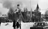 Noraskola brinner 18 februari 1966

Det brinner i en skola i Nora en vinterdag. I bildens bakgrund har brandsoldater riktat en brandspruta mot byggnadens tak för att försöka släcka elden med vatten. Grupper med skolelever står i snön på skolområdet och tittar på. Till höger i bakgrunden av bilden syns en kyrka och en lyftkran. I förgrunden av bilden står en bil med ett antal personer i. Till vänster syns en cykel som ligger i snön.