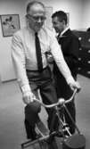 Testar spänsten 4 mars 1966

En äldre herre cyklar på en motionscykel. Han är klädd i i en ljus randig skjorta med mörk slips, gråa byxor samt mörka skor. Bredvid honom står en annan herre och lyssnar på hans hjärta med stetoskop. Han är klädd i en mörk jacka, mörka byxor, vit skjorta samt en mörk slips. I bakgrunden till vänster syns en tavla på väggen med blommor på. En annan tavla i mitten är fylld med text samt längre till höger syns ytterligare en till tavla. På golvet till höger står ett arkivskåp med många lådor i fyra våningar.