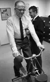 Testar spänsten 4 mars 1966

En äldre herre cyklar på en motionscykel. Han är klädd i i en ljus randig skjorta med mörk slips, gråa byxor samt mörka skor. Bredvid honom står en annan herre och lyssnar på hans hjärta med stetoskop. Han är klädd i en mörk jacka, mörka byxor, vit skjorta samt en mörk slips. I bakgrunden till vänster syns en tavla på väggen med blommor på. En annan tavla i mitten är fylld med text samt längre till höger syns ytterligare en till tavla. På golvet till höger står ett arkivskåp med många lådor i fyra våningar.