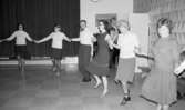 Dansskola, 15 februari 1966

Sex personer dansar i ring och håller varandra i händerna. Ytterligare andra dansar med men syns inte på bilden. Bakom dem till höger sitter en kvinna på en bänk. Längre bort syns en man som spelar dragspel.  Bakom honom står ett piano, en stol och en väska. Bredvid syns en öppen dörr. På väggen hänger en tavla. Till vänster i bakgrunden står en bänk på golvet. Över denna hänger en tjockt draperi. På golvet syns det ett mönster av kontrasterande ljusare och mörkare kvadratiska rutor. På väggen syns en mönstrad tapet.