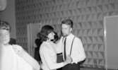 Dansskola, 15 februari 1966

I förgrunden syns två dansande par. Längst till vänster syns bara ryggtavlan på mannen medan kvinnans ansikte och vänstra arm syns tydligt. Paret till höger i bilden syns tydligt. Kvinnan är klädd i en längärmad vit blus. Hon har axellångt mörkt hår och ett armband på sin högra arm. Mannen är klädd i vit skjorta, mörka byxor, hängslen, mörk slips samt har skägg och kortklippt hår. I bakgrunden står tre andra personer. En rislampa hänger i taket och på väggarna syns en mönstrad tapet.