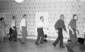 Dansskola, 15 februari 1966

Fem personer dansar i ring och håller varandra i händerna. Ytterligare personer dansar med dem men syns ej på bilden. I bakgrunden till höger syns fönster med gardiner. I mitten i bakgrunden syns en dörr samt längre till vänster ribbstolar med gymnastikmattor. På golvet syns det ett mönster av kontrasterande ljusare och mörkare kvadratiska rutor.
