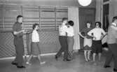 Dansskola, 15 februari 1966

Sex personer dansar i ring varav fem män och en kvinna. På väggen bakom dem syns en mönstrad tapet med en bänk stående på golvet. På och vid denna bänk ligger kläder och handväskor. En person sitter på bänken. Två dörrar syns även på bilden. På golvet syns det ett mönster av kontrasterande ljusare och mörkare kvadratiska rutor.