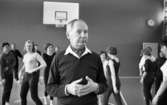 Mr Idla, 16 februari 1966

I bildens förgrund står idrottsledare Ernst Idla. Han är klädd i en mörk tröja med vit krage samt mörka byxor. I halsen har han en scarf. I bakgrunden syns flera gymnaster dansa. På väggen bakom dem hänger en basketkorg. En dörr och en klocka syns även på bilden.