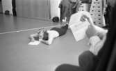 Mr Idla, 16 februari 1966

I förgrunden syns en ung flicka, en kvinnlig gymnast, som ligger på mage på golvet. Hon är klädd i en långärmad vit tröja samt svarta åtsmitande långbyxor. I håret har hon en rosett. Hon har en penna i sin hand och skriver på ett papper. Till höger syns en bit av en man som håller fram ett kuvert. I bildens bakgrund syns tre plintar. På den mittersta plinten sitter en kvinna. Vid den vänstra står en kvinna och skriver med ryggen åt kameran. En stor väska står på golvet vid hennes fötter. Vid plinten till höger står en man. Han har en kamera i handen. De tre sistnämndas huvuden har ej kommit med på bilden. Längre bort till vänster står en man vid väggen. Endast hans ben är synliga på bilden. Längre bort till vänster står en portfölj vid väggen.