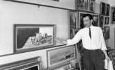 Tavlor 3 maj 1966

Man som håller i ett pappersblock och en penna, står vid tavla och förevisar med handen en tavla med stillebenmotiv