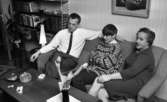 Brudpar 1946 (hemma hos), 9 mars 1966

En familj sitter i en soffa i ett vardagsrum. Familjen består av far, mor samt en ung son i nedre tonåren. På ett bord framför mannen i huset ligger ett paket cigaretter. På väggarna hänger tavlor. Mannen röker.