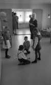 Barndaghem 29 mars 1966

I förgrunden syns det två pojkar som sitter på golvet och leker med pusselbitar av trä. En flicka syns det stående bredvid dem till höger och en annan flicka till vänster. Pojkar har kort klippta frisyrer. Flickan till höger har toffsar och är klädd i strumpbyxor och klännning, samt mörkare skor. Flicka till vänster är klädd i strumpbyxor, kjol, jacka, och har kortklippt frisyr. Båda pojkar är klädda i mörka byxor och en ljusare skjorta med flyga. En av pojkarna har en pullover på sig och en annan pojke har hängslen. Den kvinnliga lärare bakom är klädd i en klänning med en ljusare krage och skor.
 I bakgrunden syns det borden som är i storleken anpassade för barn. Borden är dukade med bland annat kaffe koppar och sockerskål.