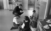 Barndaghem 29 mars 1966

I förgrunden syns det två flickor och två pojkar som sitter på golvet och leker med pusselbitar av trä. En man som har en kortklippt frisyr, en ring på den högra handens fingret, bär glassögon och som är klädd i mörk kavaj, byxor och skor [...]