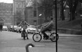 Mopedister 5 maj 1966

Två unga pojkar åker på varsin moped på en gata i centrala Örebro. De skrattar. De är klädda i jackor, byxor och skor. I bakgrunden syns bilar som står parkerade, byggnader samt en man i mörka kläder som går på gatan med ryggen åt kameran.