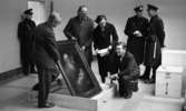 Nationalmuseum 1 31 maj 1966

Två män håller upp en tavla föreställande Simeon och Jesusbarnet. De håller på att packa upp den ur en låda som ligger på golvet. En av männen är klädd i ljus kostym och den andre mannen bär arbetsrock. En man i svart kostym står i knäböjande ställning nära tavlan på golvet. En kvinna i svart kappa står bredvid honom och håller ett papper i sina händer. Tre stycken polismän står i rummet även. Ytterligare en man syns delvis bakom mannen i arbetsrock.