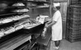Henning Kjellgrens bageri,18 april 1966

En kvinna i vita arbetskläder samt vit hatt på huvudet håller i en påse med vänstra handen. I högra handen håller hon i ett ljust, avlångt bröd som håller på att lägga i påsen. En kartong med texten 