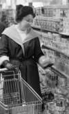 Burkmat 24 februari 1966

En kvinna klädd i mössa med tofs, kappa, tröja samt handskar betraktar en konservburk som hon håller i sin vänstra hand. Hon har en kundvagn med sig. Hennes handväska samt varor står i kundvagnen. Hon står intill hyllor fyllda med varor: paket samt konservburkar.