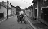 Noramarknad, Simmar i Gustavsvik 4 september 1965 

Tre äldre damer går arm i arm på en gata i Nora. De är klädda i kappor, två av dem bär hattar och alla bär handväskor i sina händer. Byggnader syns i bakgrunden.