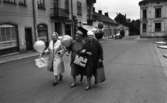 Noramarknad, Simmar i Gustavsvik 4 september 1965 

Tre äldre damer går på en gata i Nora. De är klädda i kappor, två av dem bär hattar och alla bär handväskor i sina händer. De har även var sin ballong i händerna. I bakgrunden syns byggnader samt en parkerad bil.