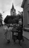 Noramarknad, Simmar i Gustavsvik 4 september 1965 

Tre äldre damer promenerar arm i arm på en gata i Nora. Alla tre är klädda i kappor, bär handväskor och varsin ballong i händerna. Två av dem har hatt på huvudet. En kyrka och andra byggnader syns i bakgrunden. En cyklist samt flera parkerade bilar syns även.