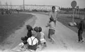 Oläslig rubrik 23 augusti 1965
 
I förgrunden syns fem barn som sitter på en fyrkantig bräda. I ena änden av brädan sitter ett snöre. Snöret har en ring i ena änden som hålls av en vuxen man som drar barnens bräda med hjälp av snöret. En pojke hjälper till medan en annan pojke går bredvid. De befinner sig på en grusväg. I bakgrunden syns ett bostadsområde med bilparkering framför.