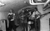 Oläslig rubrik 23 augusti 1965
 
Ett rockband spelar i en källarlokal. I förgrunden står tre unga pojkar och spelar på varsin gitarr. I bakgrunden sitter ytterligare en pojke och spelar trummor. Till vänster står en högtalare. Ett tygstycke hänger på väggen bakom trummisen.