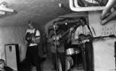 Oläslig rubrik 23 augusti 1965
 
Tre tonårspojkar spelar gitarr i en källarlokal. I bakgrunden sitter ytterligare en pojke och spelar trummor. En pojke sitter på golvet och lyssnar invid en högtalare.