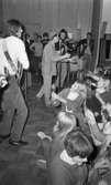 Popgala i Kumla, 24 augusti 1965

Ett rockband spelar på en scen i Kumla. En gitarrist och en tamburinspelare syns på bilden. Mellan dem står en sångare i en Sherlock Holmesinspirerad hatt och sjunger i en mikrofon. Ett piano syns i bakgrunden. Nedanför scenen står publiken. En tonårsflicka i publiken spelar på en marackas.