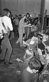 Popgala i Kumla, 24 augusti 1965

Ett rockband spelar på en scen i Kumla. En gitarrist och en tamburinspelare syns på bilden. Mellan dem står en sångare i en Sherlock Holmesinspirerad hatt och sjunger i en mikrofon. Ett piano syns i bakgrunden. Nedanför scenen står publiken. En tonårsflicka i publiken spelar på en marackas.