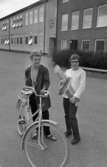 Vivalla problem, Trafiktävling, Konfirmation 10 maj 1966

Två unga tonårsflickor står på en skolgård. Flickan till vänster har kort hår och håller i en cykel. Hon är klädd i jacka, byxor och tröja och har ett halsband med en klocka på om halsen. Flickan till höger är klädd i vit kavaj, vit blus och mörka byxor. Hon håller i ett avlångt plastpaket. De står framför en skolbyggnad.