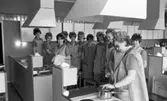 Yrkesskolan Älvtomta 25 april 1966

Kvinnor som går en yrkesutbildning i Älvtomta som berör ämnesområdet hushållskunskap står i ett kök. De är klädda i arbetskläder samt har likadana mössor på sina huvuden. De står samlade kring en spis samt en diskbänk. En av kvinnorna lyfter på locket till en kastrull och de andra betraktar detta.