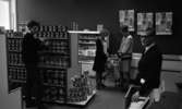 Yrkesskolan Älvtomta 25 april 1966

Inne i en affär står två män och två kvinnor. Hyllorna är fyllda av matvaror. Mannen till vänster håller på och plockar bland hyllorna medan mannen till höger står vid kassaapparaten. Han är klädd i kostym. De två kvinnorna står med en varukorg emellan sig och verkar vara i färd med att fylla en fast nätkorg med konservburkar. I bakgrunden står en frysbox vid väggen.