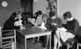 Yrkesskolan Älvtomta 25 april 1966

Sju unga kvinnor som är elever vid en yrkesskola i Älvtomta sitter tillsammans runt ett fyrkantigt bord. Två av eleverna broderar dukar, en virkar, en läser en bok, en läser en tidning, en tittar in i kameran och en skriver i ett block som hon har lagt framför sig på bordet.