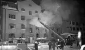 Branden under natten 9 februari 1966

En byggnad brinner och rök väller ut ur den. I förgrunden syns fem män varav två är brandmän. De står på den snöklädda marken. Mot byggnadens husfasad står tre brandmän på stegar. De sprutar vatten på huset med hjälp av brandslangar som är förankrade i två stycken brandbilar.