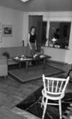 Byggspecial Oxhagen 11 februari 1966

I ett vardagsrum i en lägenhet i Oxhagen står en kvinna klädd i svart linne, grå kjol och sandaler och invid ett bord. I rummet finns en soffa, en fåtölj och en matta under bordet. I förgrunden står en vit stol samt en matta under den. Tavlor hänger på väggarna.