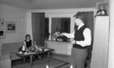 Byggspecial Oxhagen 11 februari 1966

I ett vardagsrum i Oxhagen sitter en kvinna i en soffa. Hon är klädd i svart linne, grå kjol och har sandaler på fötterna. En man står upp i förgrunden till höger invid en bokhylla. Han är klädd i vit skjorta, svart väst, svart slips och grå byxor. Han håller en bok i sin högra hand och håller upp den mot kvinnan. Uppe på bokhyllan ligger en pickelhuva. I bakgrunden syns fönster med gardiner. På väggarna hänger det tavlor.