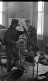 Rycket in i lumpen 8 februari 1966

Tre soldater står inne i en stor sal och klär på sig militärkläder och militärutrustning. Soldaten i förgrunden håller ett gevär i vänster hand. Militärutrustning ligger på en stol framför honom samt på golvet.