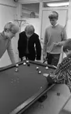 Biljard fritidssysselsättning 1 februari 1966

Fyra unga pojkar är i färd med att spela biljard i ett rum. Tre av dem är klädda i tröjor och en av dem har en kavaj på sig. En klädhängare står i bakgrunden. I bakgrunden skymtar en pojkes ansikte. Han befinner sig utanför rummet och kikar in genom ett fönster.