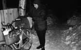 Tidningsbud 12 februari 1966

Ett tidningsbud, en kvinna, klädd i kappa och mössa binder fast en tidningsbunt på pakethållaren till en damcykel. Tidningsbunten är försedd med ett skyddshölje. På cykelstyret hänger en stor handväska. Ett par vantar samt nycklar skymtar i ytterfacket. På marken ligger en massa snö. Ett staket och ett träd syns i bakgrunden.