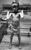 Tyngdlyftarreportage 31 januari 1966 

En pojke i åttaårsåldern med bar överkropp samt klädd i vita små shorts lyfter en skivstång upp mot axlarn inne i en träningslokal. Han sysslar med tyngdlyftning. I bakgrunden syns fyra skivstänger som ligger på golvet.