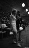 Cykel, popgala 6 juni 1966

Närbild på två unga flickor. Flickan till höger är klädd i svart klänning, svartvita lågklackade pumps och bär en vit handväska i sina händer. Flickan till vänster har vit ärmlös blus, vit kjol, vita stövlar samt bär en vit handväska i sin högra hand. I bakgrunden syns publik sitta på sina platser.