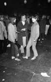 Popgala 6 juni 1966

Närbild på tre flickor ur publiken under en musikföreställning klädda i blusar, tröjor och byxor. Flickan i mitten bär kappa. Två av flickorna är barfota. Flickan till höger håller snörena till ett par sandaler i sin högra hand.